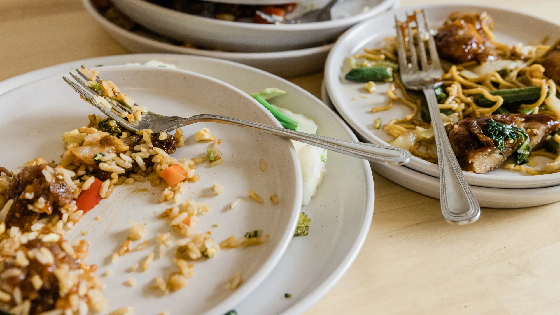 Piatti in ceramica bianca con avanzi di cibo, riso, verdure e noodles sparsi, e forchette lasciate sopra, a rappresentare lo spreco alimentare post pasto.