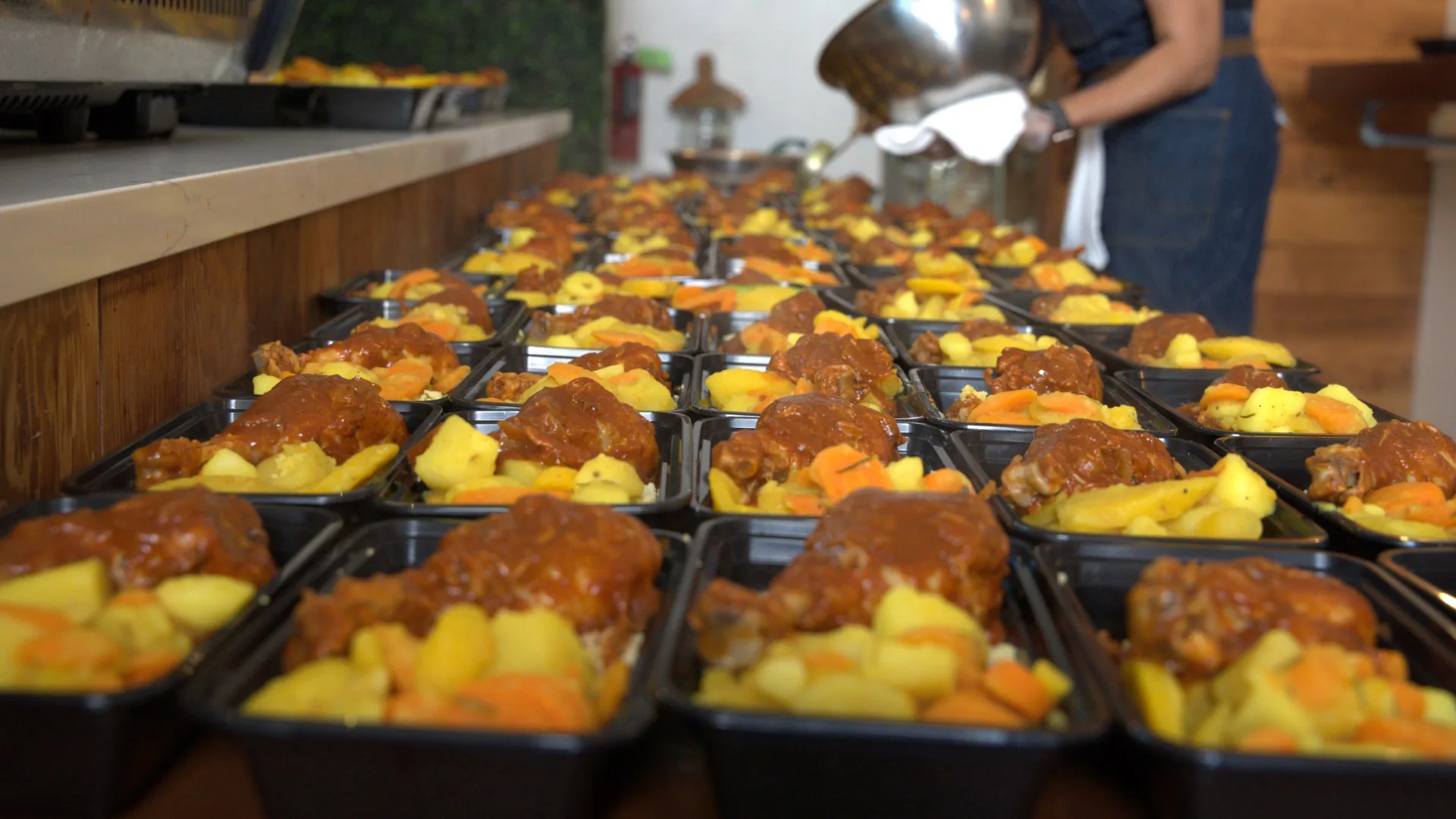 A long row of black single-portion trays with ready meals featuring chicken in sauce, potatoes, and carrots, arranged on a work surface in an industrial kitchen, with a person in the background serving the food.
